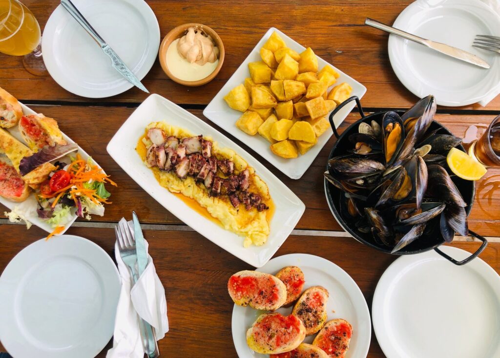 Mesa de madera con varios platos para compartir: patatas, mejillones, pulpo y pan con tomate